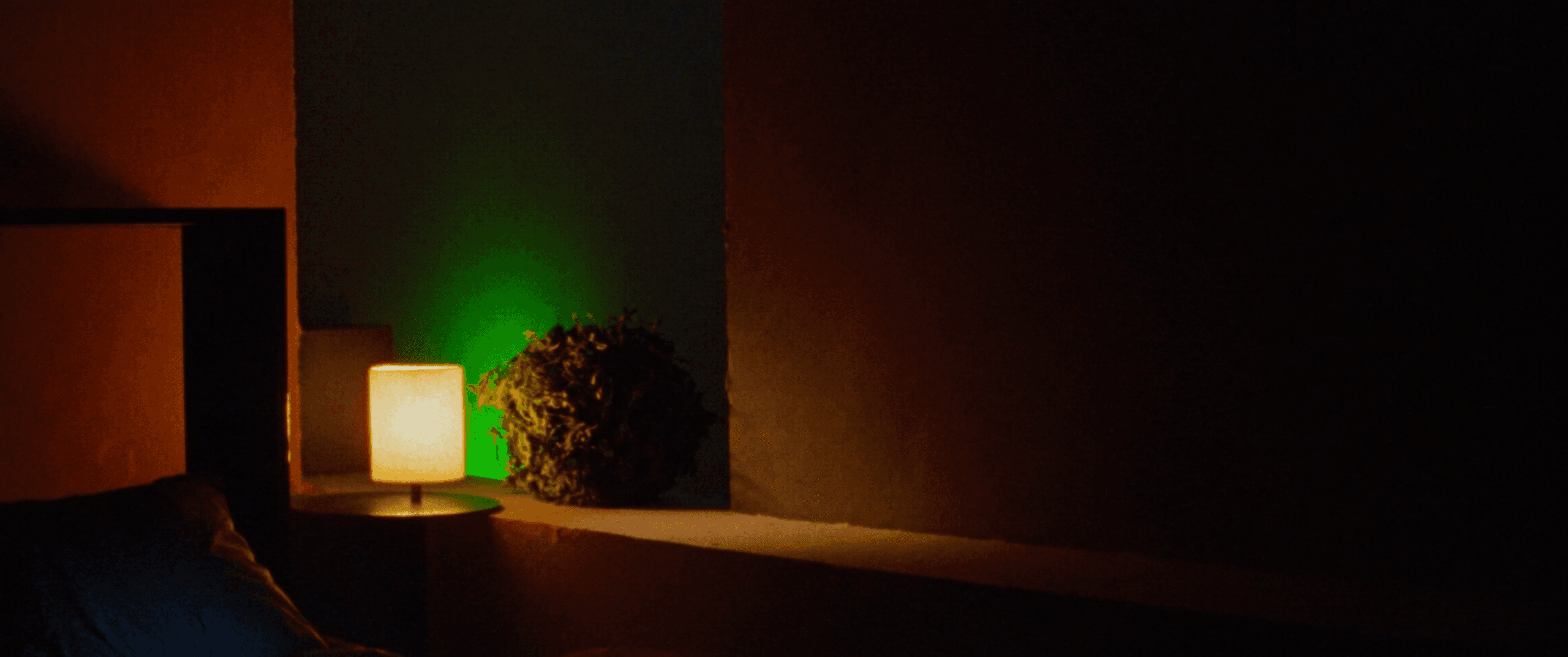 A cozy, dark bedroom corner at night with a warm lamp glow and a plant lit by an intense green accent light
